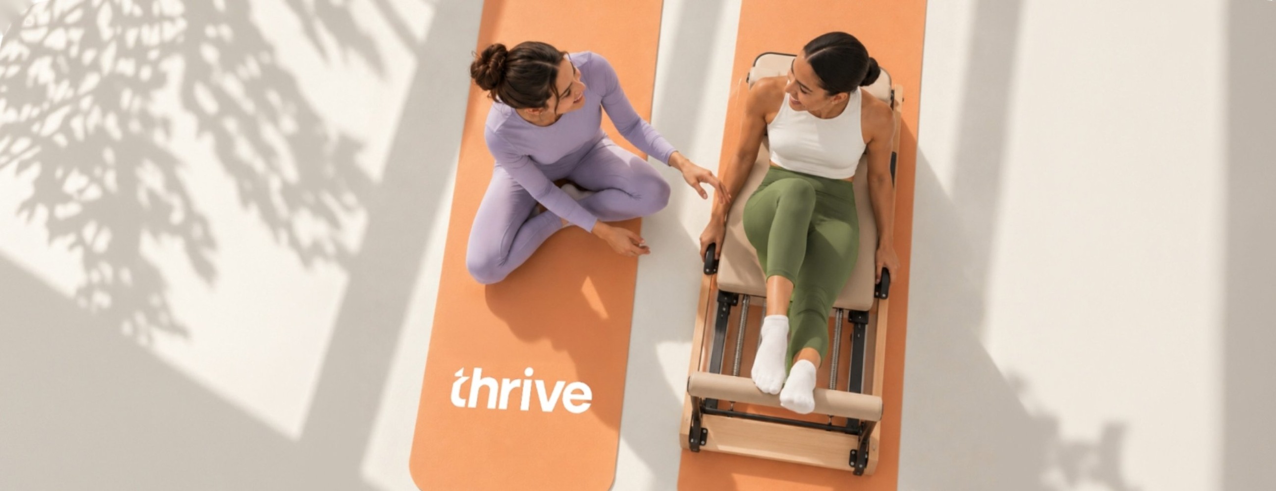 Two women on orange Thrive mats, one on a pilates reformer, chatting in a sunlit studio